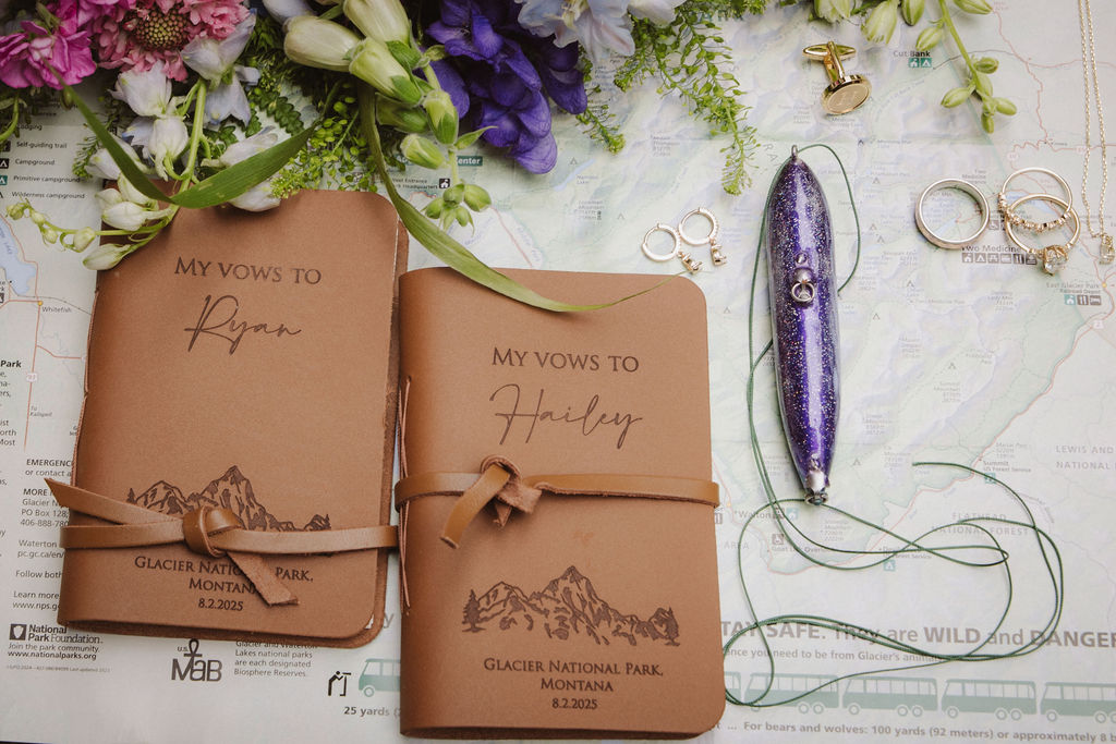 Two brown vow books labeled "My Vows to Ryan" and "My Vows to Hailey" surrounded by rings, a purple pen, and flowers on a map background.