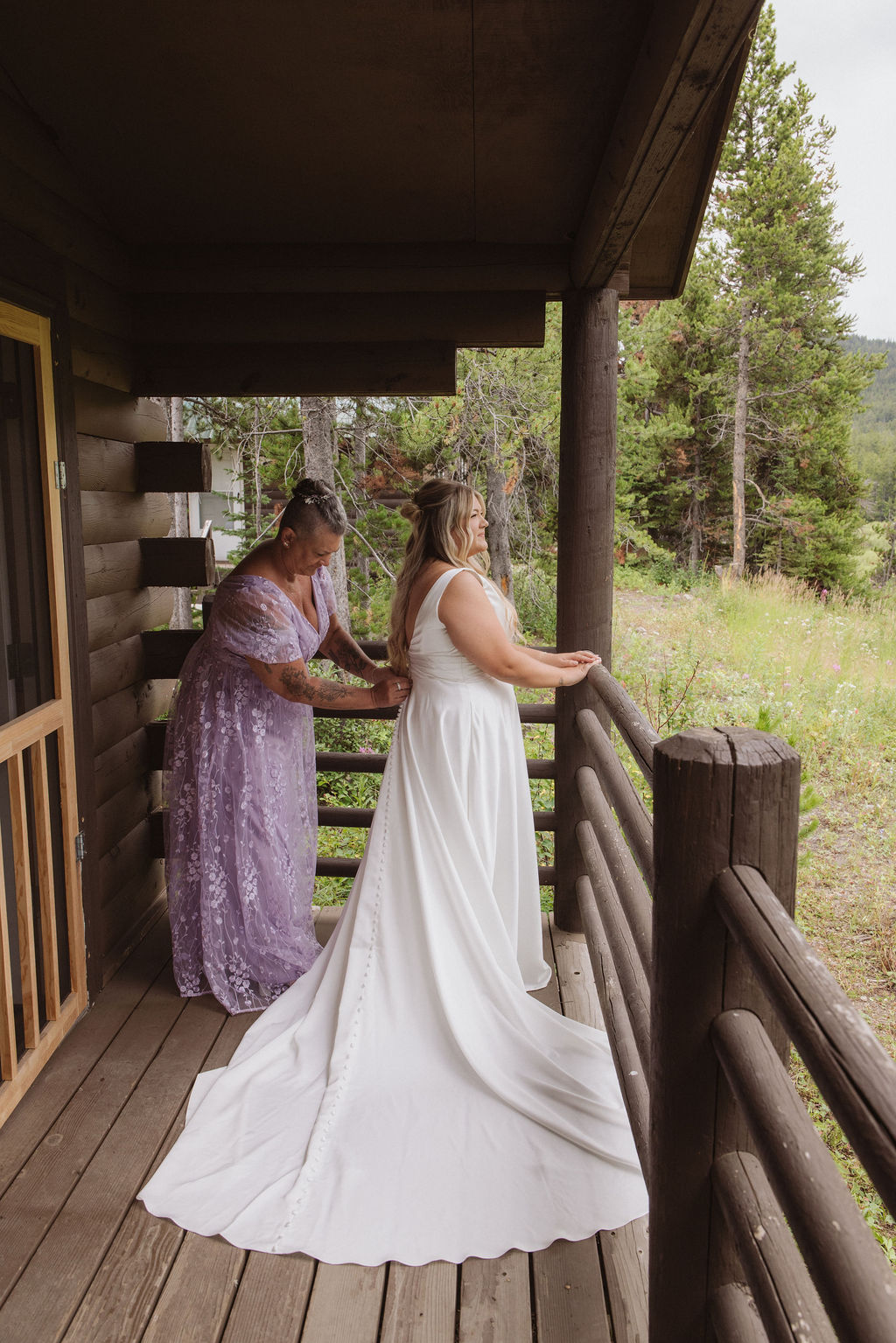 A woman in a lavender dress helps a bride in a white gown with her dress on a rustic wooden porch overlooking a grassy landscape.