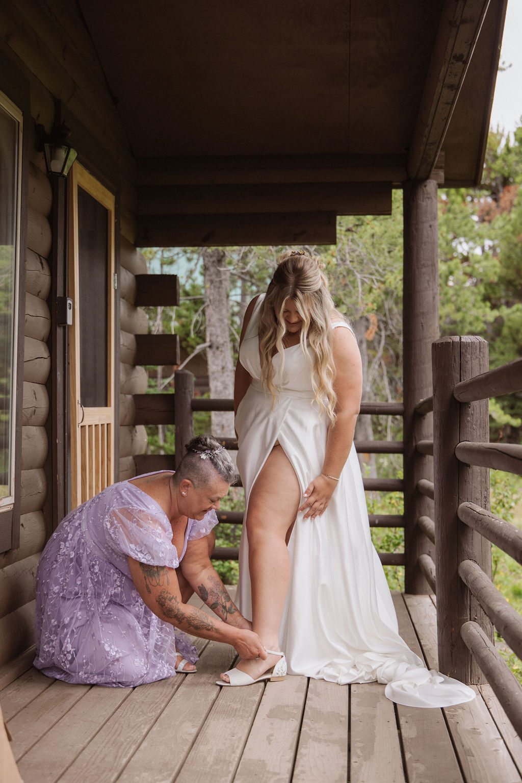 A woman in a lavender dress helps a bride in a white gown with her dress on a rustic wooden porch overlooking a grassy landscape.