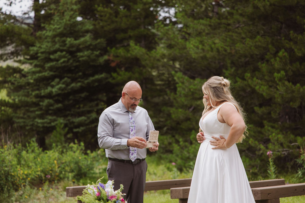 A man in a dress shirt reads from a notecard while a woman in a white wedding dress stands nearby outdoors, surrounded by greenery.