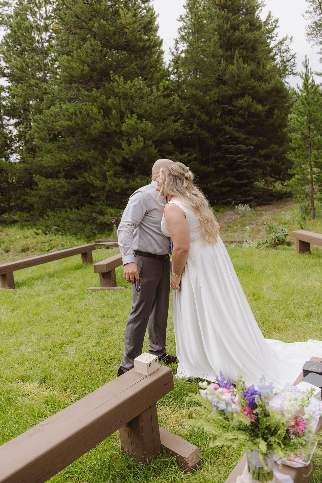 A bride and groom kiss outdoors in a grassy area surrounded by wooden benches and trees, with a bouquet of flowers visible in the foreground.