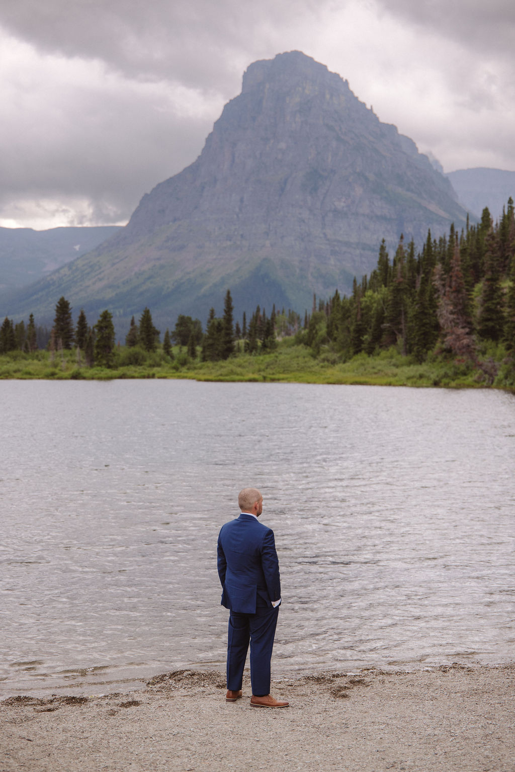 A man in a blue suit stands on the shore of a lake, facing the water, with a large mountain and cloudy sky in the background.