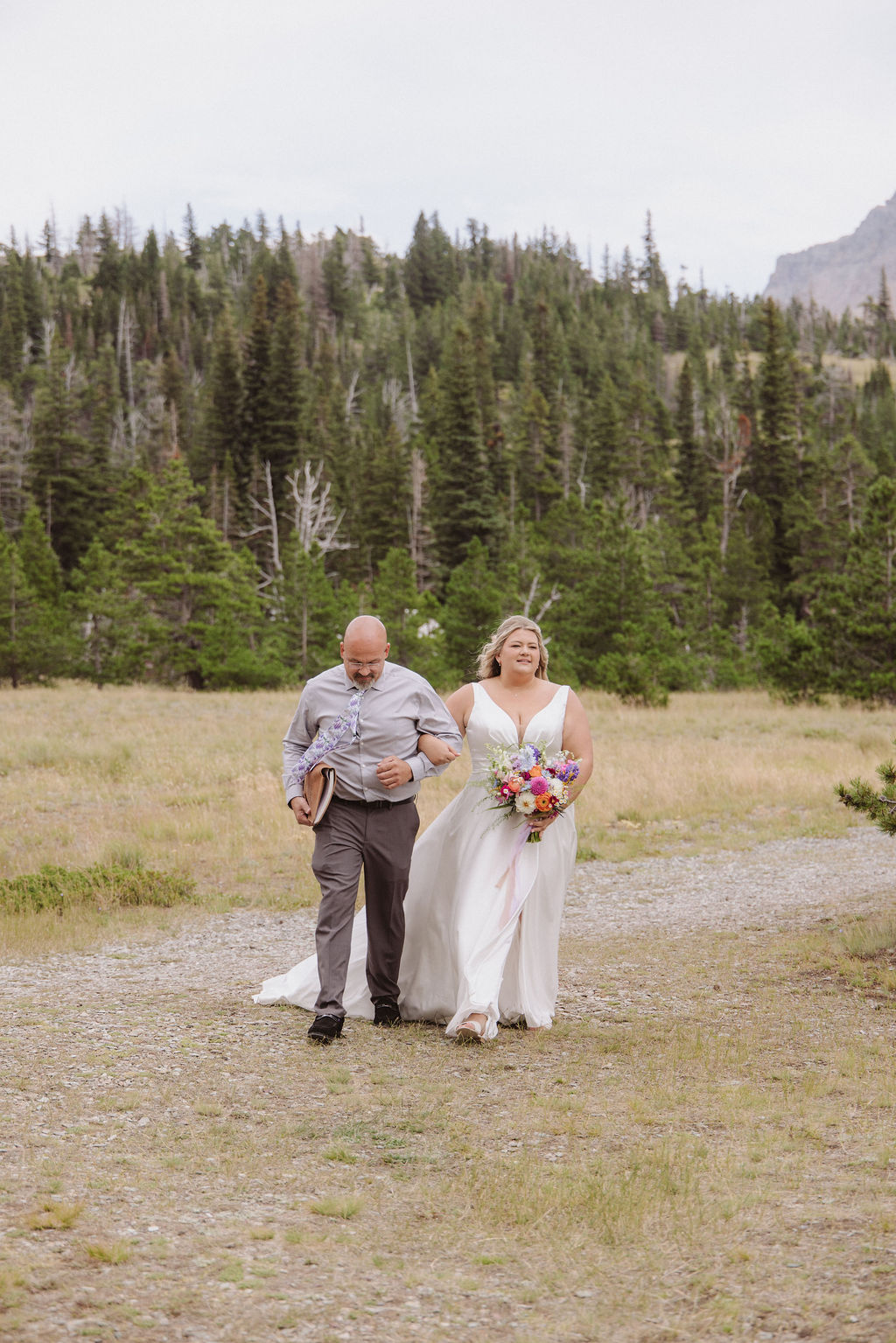 A bride in a white dress holding a bouquet walks beside a man on a gravel path, with trees and mountains in the background.