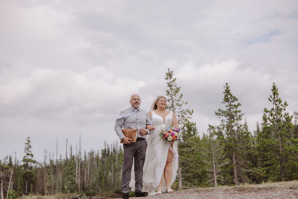A bride in a white dress holding a bouquet walks outdoors beside a man in a shirt and tie, with trees and a cloudy sky in the background.