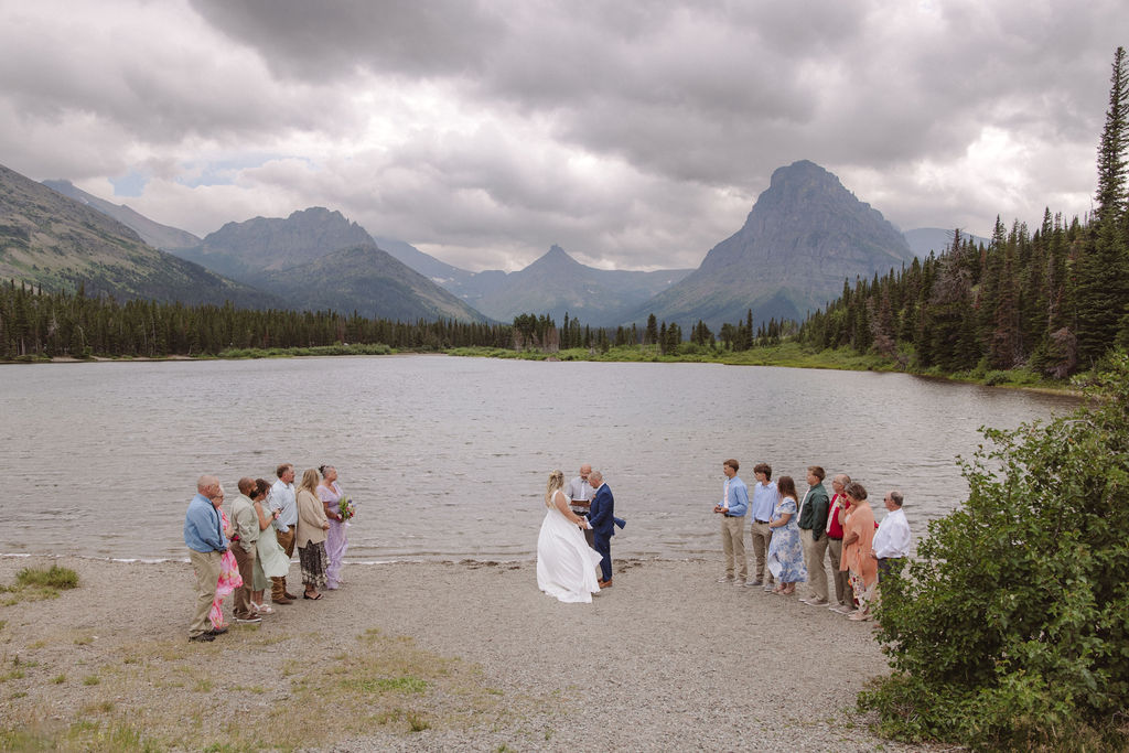 A wedding ceremony is taking place on a lakeshore with mountains and clouds in the background, attended by a small group of people standing in two lines.