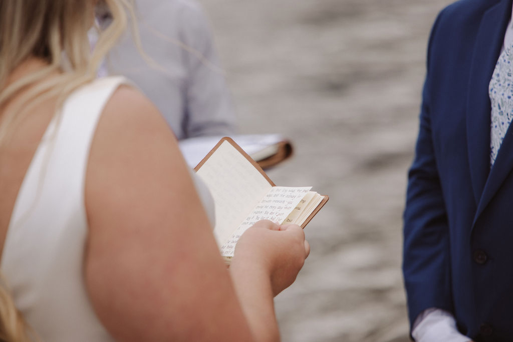 A person in a white dress holds an open notebook and reads during an outdoor ceremony, facing another person in a blue suit.