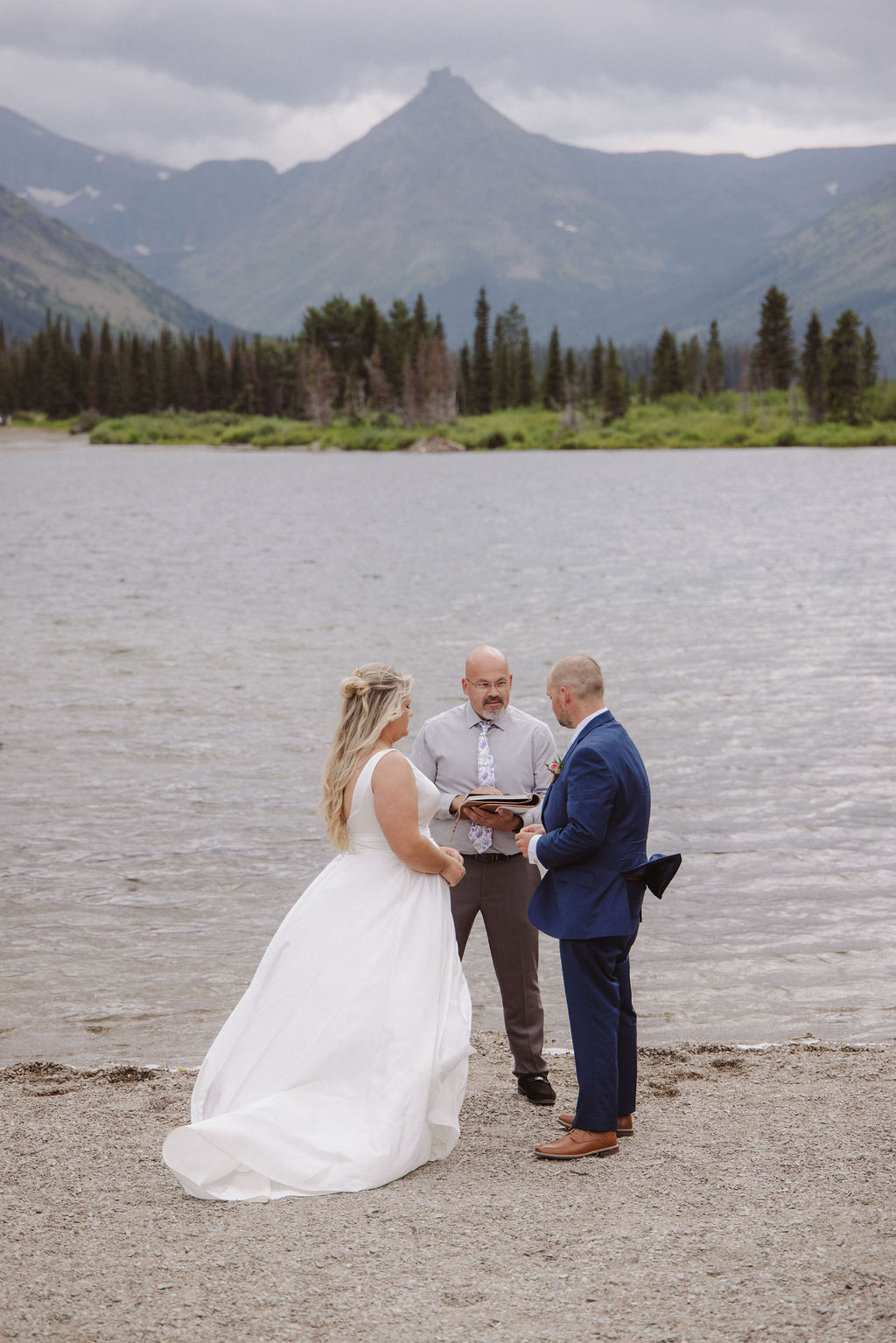 A bride and groom stand with an officiant on a lakeshore, with mountains and trees in the background under a cloudy sky.