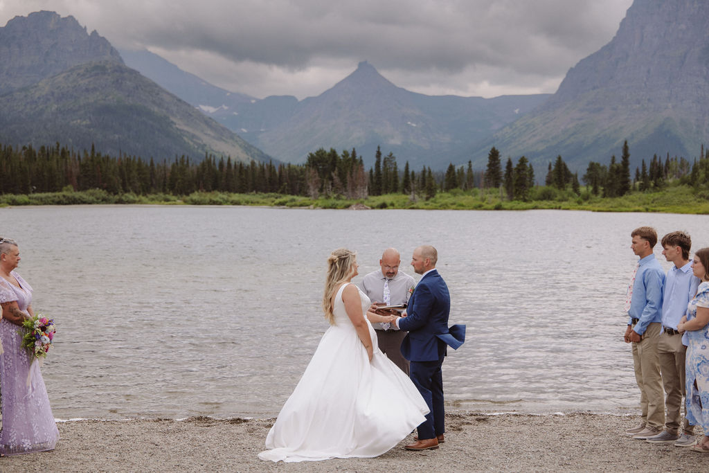 A wedding ceremony is taking place on a lakeshore with mountains and clouds in the background, attended by a small group of people standing in two lines.