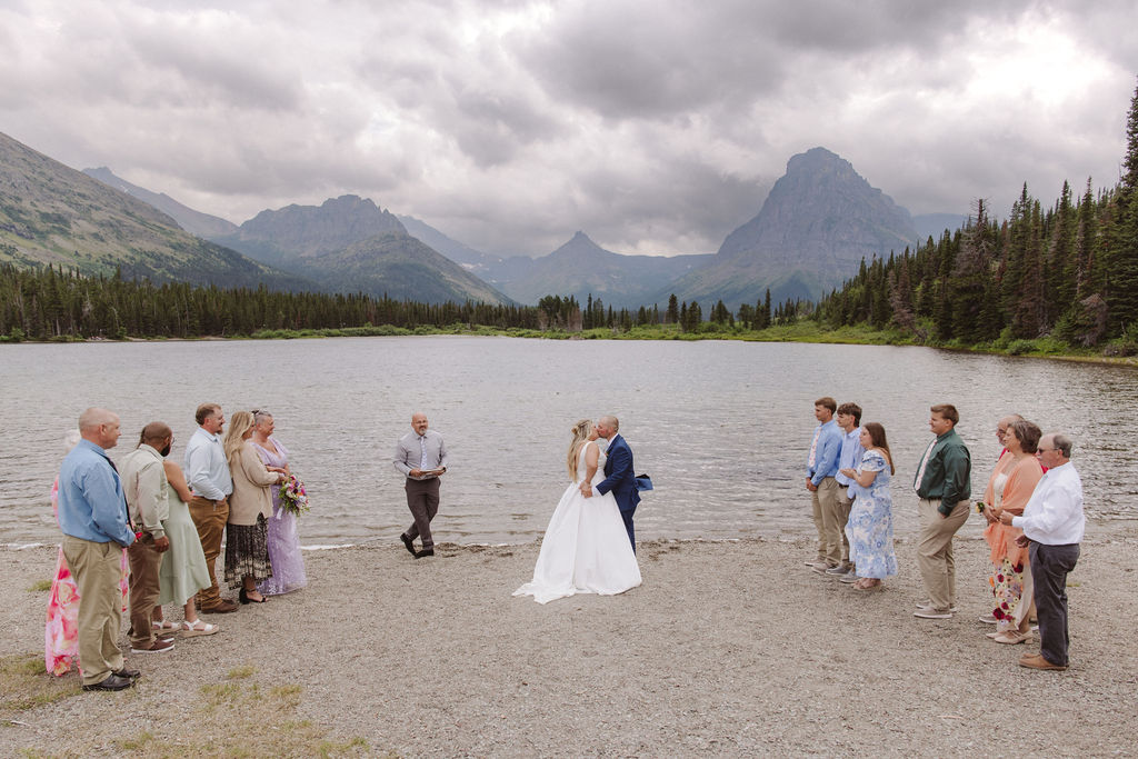 A wedding ceremony is taking place on a lakeshore with mountains and clouds in the background, attended by a small group of people standing in two lines.