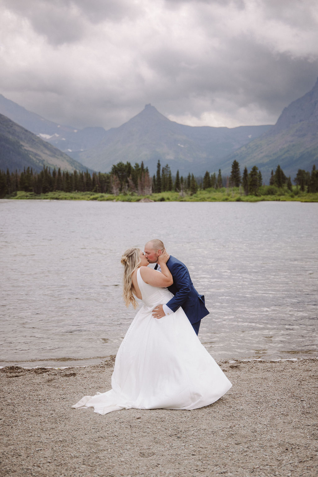 A wedding ceremony is taking place on a lakeshore with mountains and clouds in the background, attended by a small group of people standing in two lines.