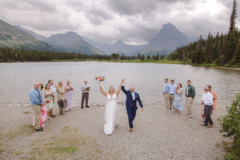 A wedding ceremony is taking place on a lakeshore with mountains and clouds in the background, attended by a small group of people standing in two lines.