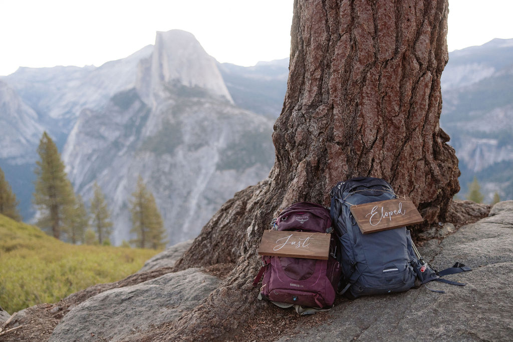 Two backpacks rest against a large tree with wooden signs attached; mountains and trees are visible in the background for a Yosemite elopement