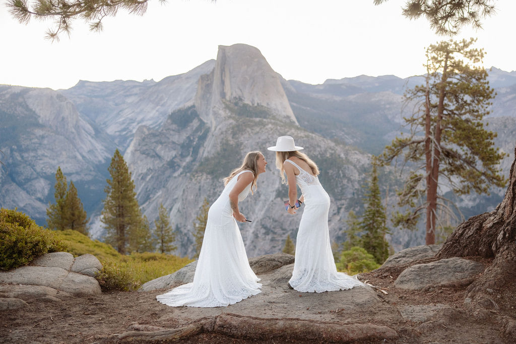 Two women in white dresses stand facing each other and smiling on a rocky outcrop, with a mountain and trees in the background.