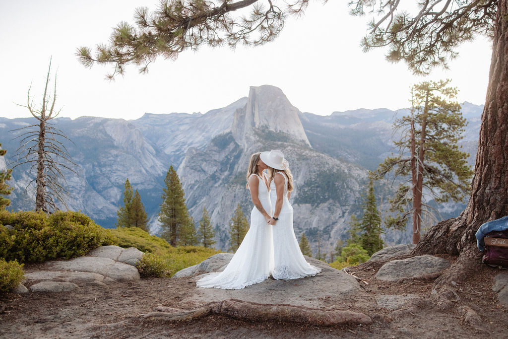 Two women in white dresses stand facing each other and smiling on a rocky outcrop, with a mountain and trees in the background.