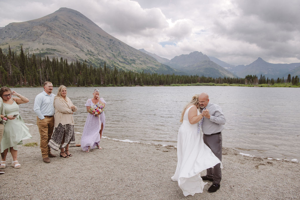 A wedding ceremony is taking place on a lakeshore with mountains and clouds in the background, attended by a small group of people standing in two lines.