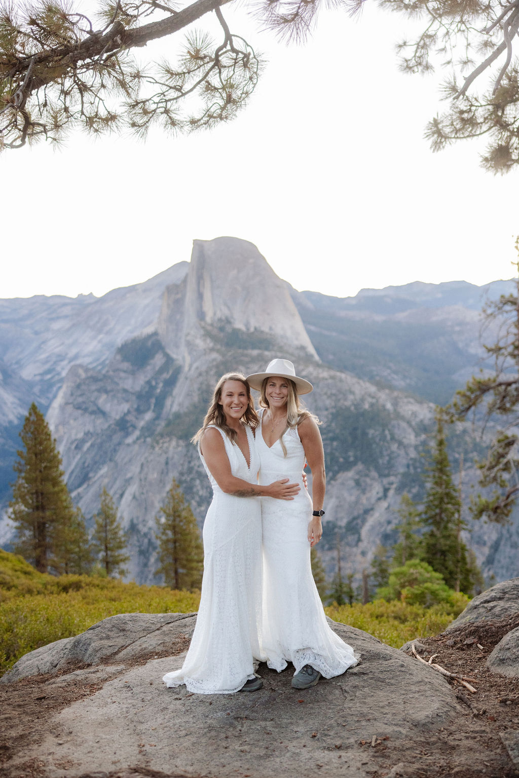 Two women in white dresses stand facing each other and smiling on a rocky outcrop, with a mountain and trees in the background.