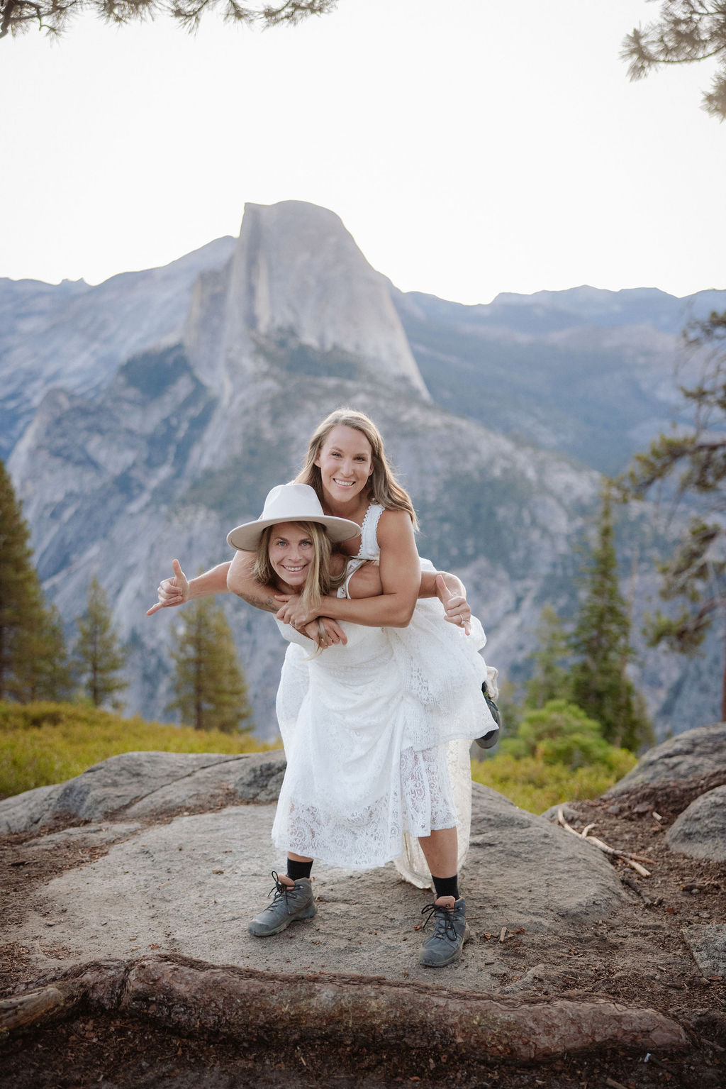 Two women in white dresses stand facing each other and smiling on a rocky outcrop, with a mountain and trees in the background.