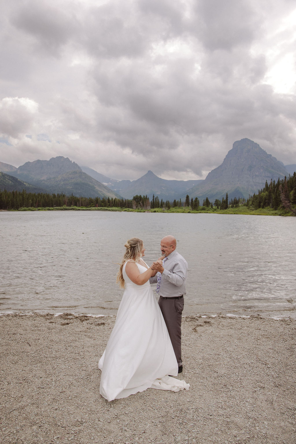 A wedding ceremony is taking place on a lakeshore with mountains and clouds in the background, attended by a small group of people standing in two lines.