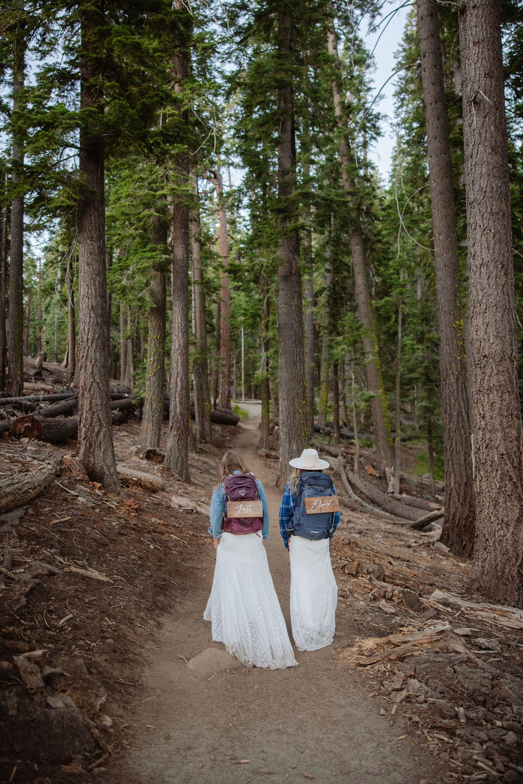 Two people wearing white dresses, hats, and backpacks walk along a forest trail surrounded by tall trees.