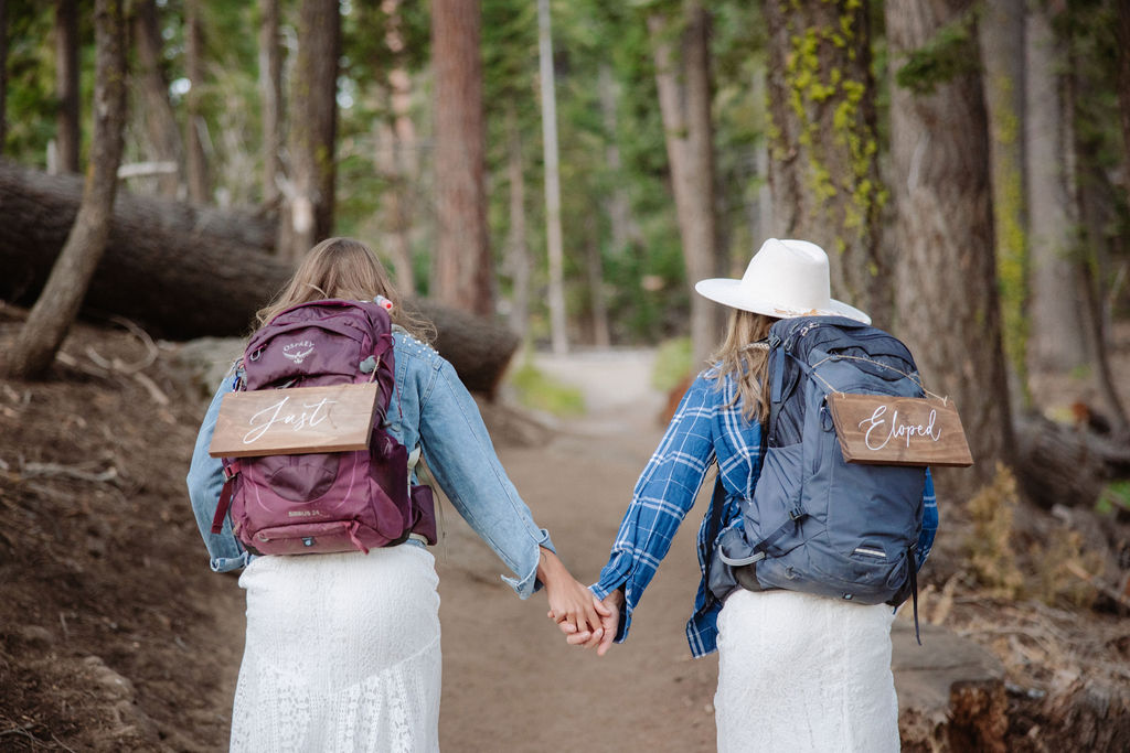 Two people wearing white dresses, hats, and backpacks walk along a forest trail surrounded by tall trees.
