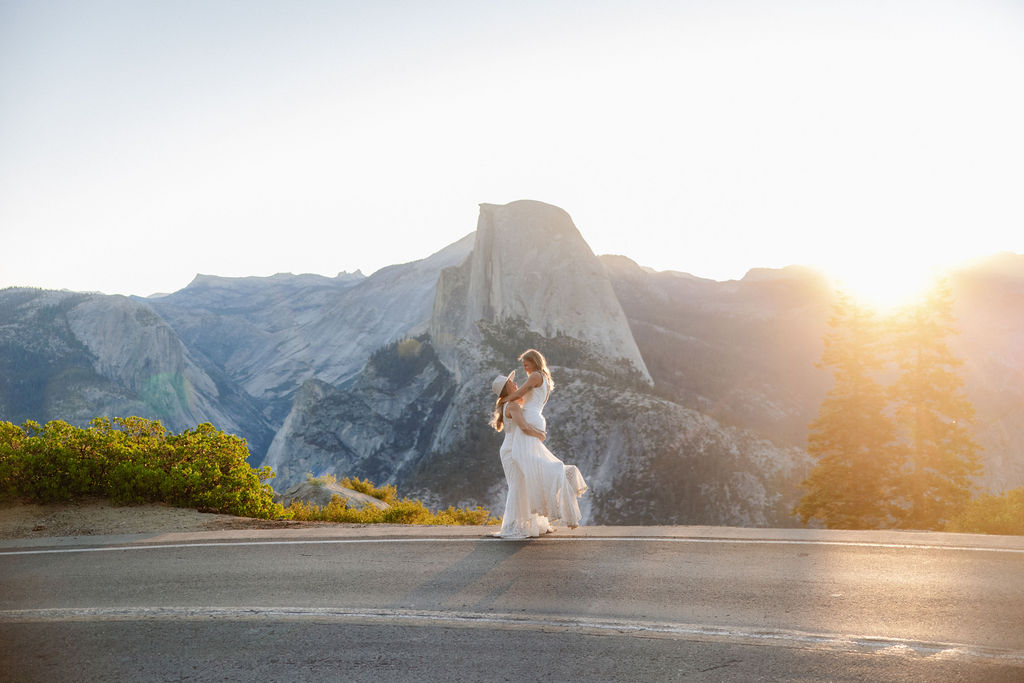 A bride and groom embrace on a roadside with mountains and the sun setting behind them.