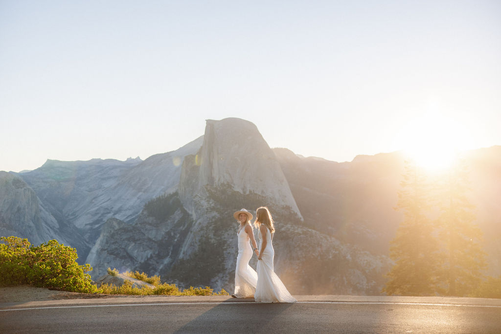 A bride and groom embrace on a roadside with mountains and the sun setting behind them.