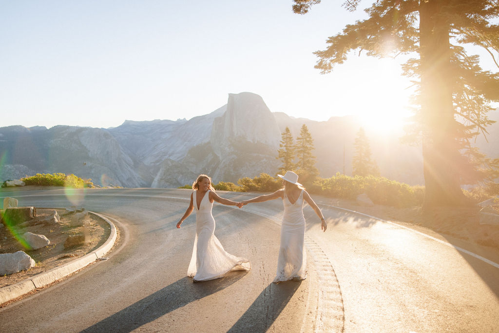 A bride and groom embrace on a roadside with mountains and the sun setting behind them.
