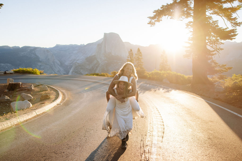A bride and groom embrace on a roadside with mountains and the sun setting behind them.