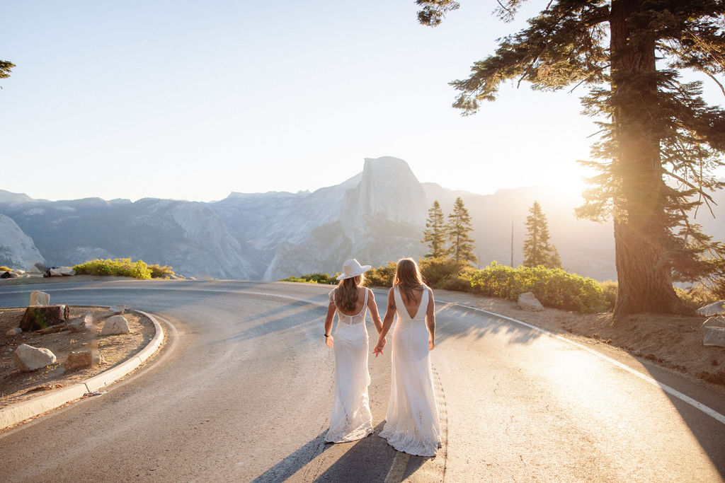 A bride and groom embrace on a roadside with mountains and the sun setting behind them.