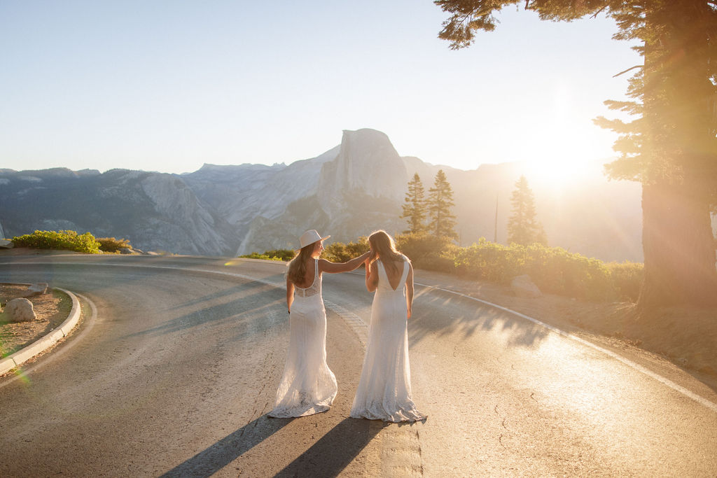 Two people in white dresses stand on a curved road at sunset, surrounded by mountains and trees, holding hands and facing away from the camera.