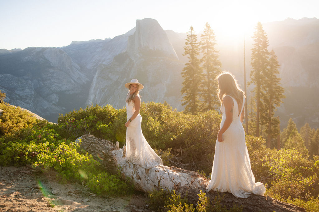 Two women in white dresses stand on a fallen log in a sunlit forest with mountains in the background for a Yosemite elopement