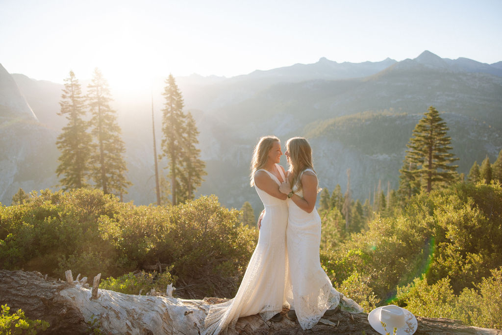 Two women in white dresses stand on a fallen log in a sunlit forest with mountains in the background for a Yosemite elopement