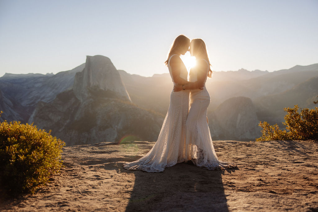 Two women in white dresses stand on a fallen log in a sunlit forest with mountains in the background for a Yosemite elopement