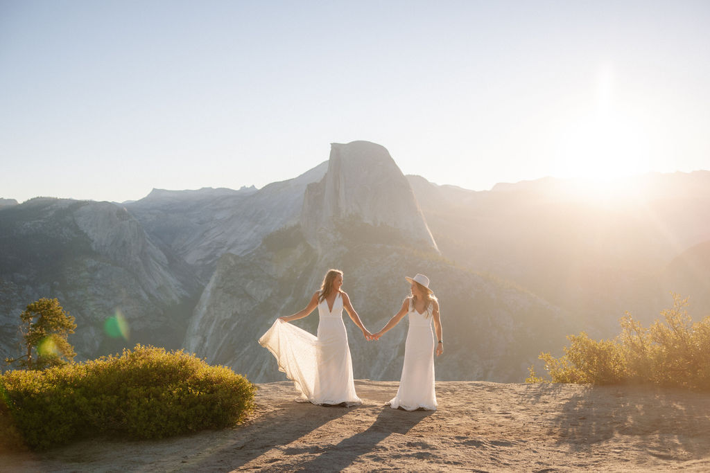 Two women in white dresses stand on a fallen log in a sunlit forest with mountains in the background for a Yosemite elopement