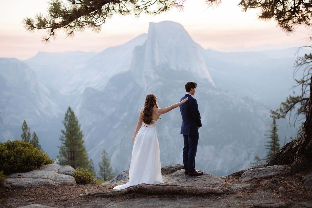 A bride in a white dress stands beside a groom in a suit on a rocky ledge, with mountains and trees in the background for a wedding at glacier point