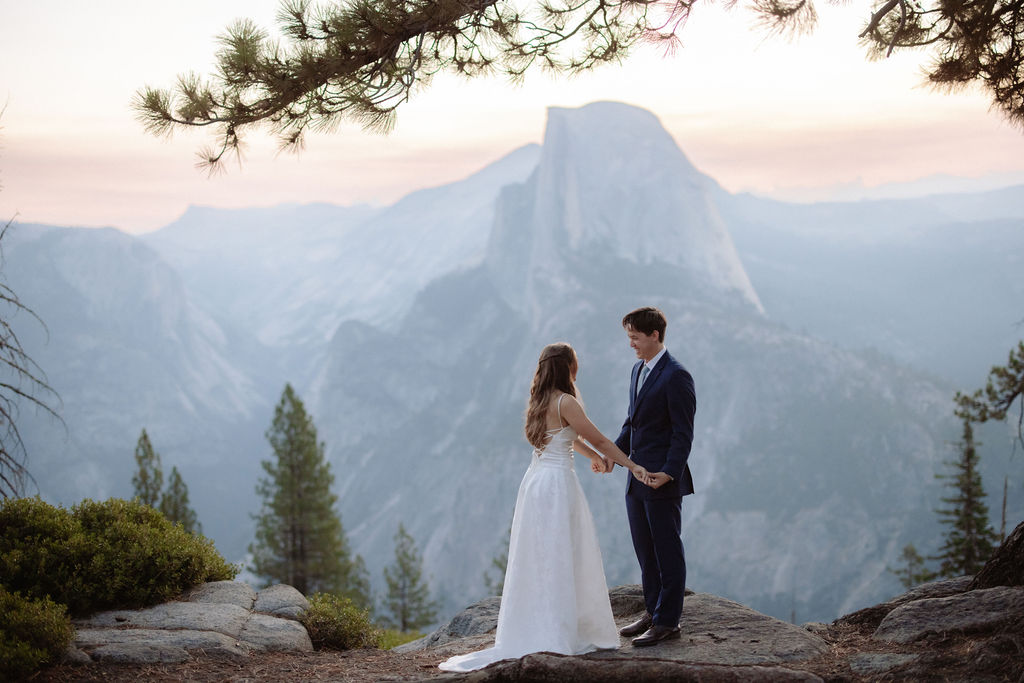 A bride in a white dress stands beside a groom in a suit on a rocky ledge, with mountains and trees in the background for a wedding at glacier point