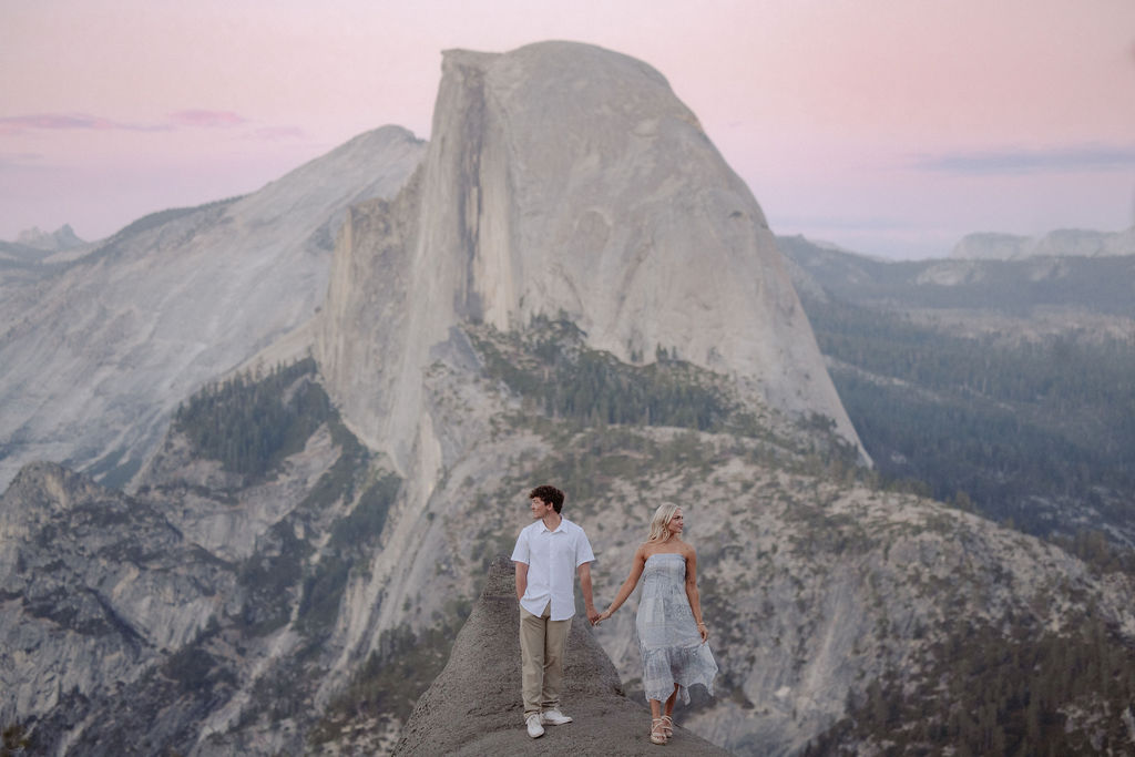 A couple holding hands stands on a rocky ledge overlooking Half Dome at Yosemite National Park during sunset.