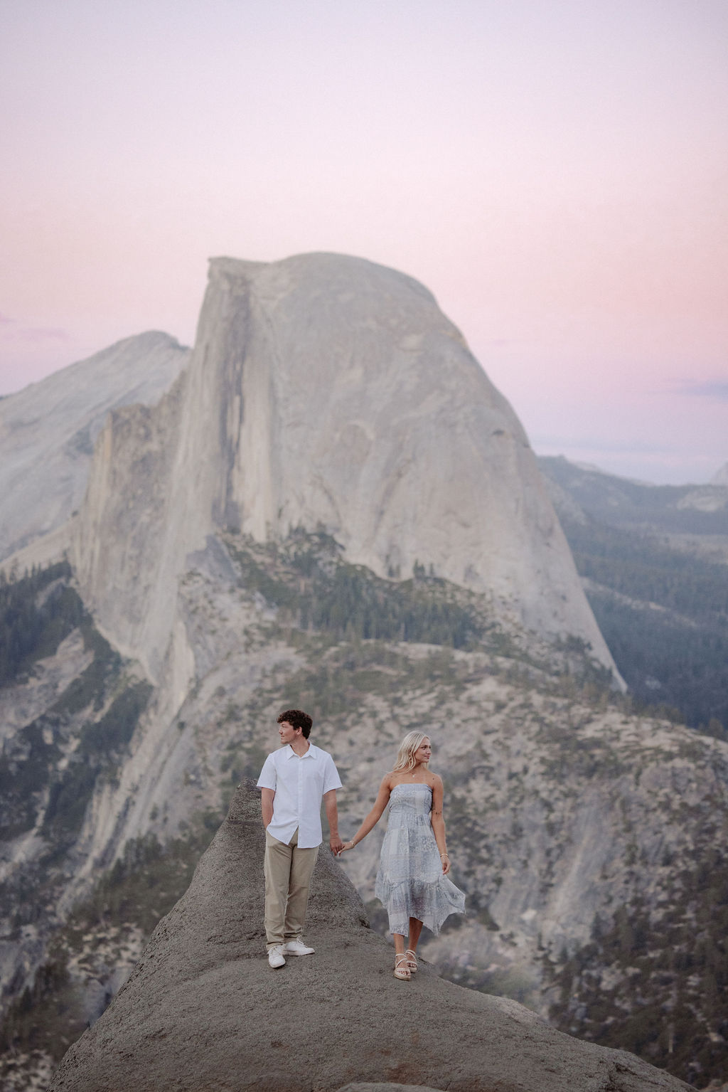 A couple holding hands stands on a rocky ledge overlooking Half Dome at Yosemite National Park during sunset.