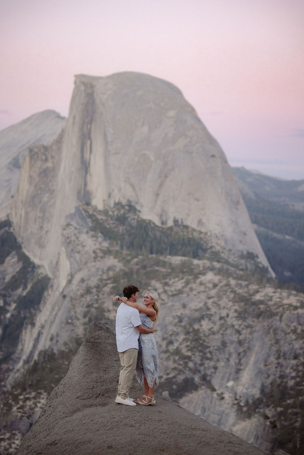 A couple holding hands stands on a rocky ledge overlooking Half Dome at Yosemite National Park during sunset.