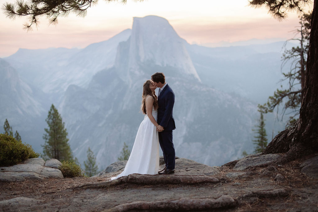 A bride in a white dress stands beside a groom in a suit on a rocky ledge, with mountains and trees in the background for a wedding at glacier point