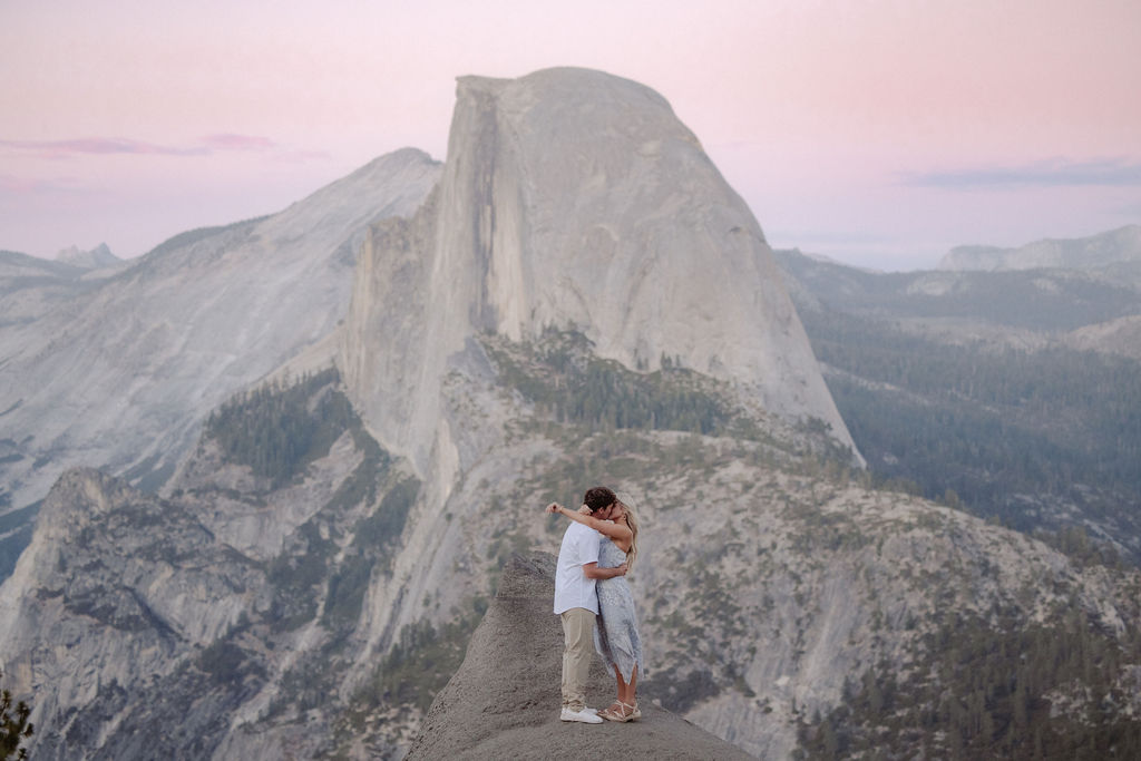 A couple holding hands stands on a rocky ledge overlooking Half Dome at Yosemite National Park during sunset.