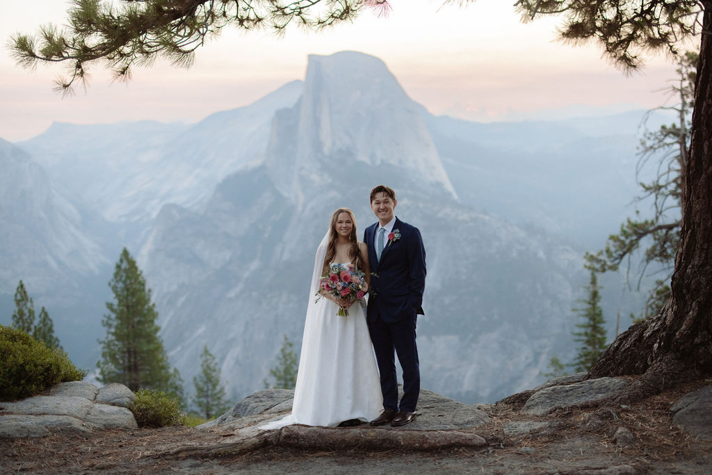 A bride in a white dress stands beside a groom in a suit on a rocky ledge, with mountains and trees in the background for a wedding at glacier point