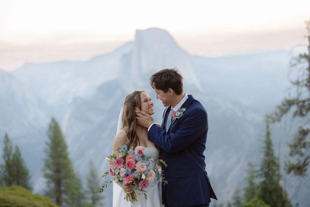 A bride in a white dress stands beside a groom in a suit on a rocky ledge, with mountains and trees in the background for a wedding at glacier point