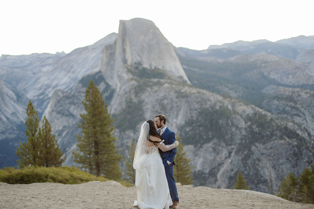A bride and groom embrace outdoors with mountains and trees in the background, including a prominent granite peak for a wedding at yosemite