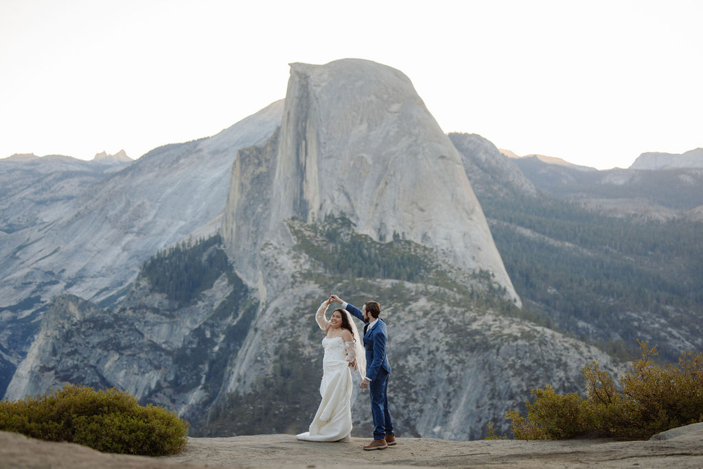 A bride and groom embrace outdoors with mountains and trees in the background, including a prominent granite peak for a wedding at yosemite