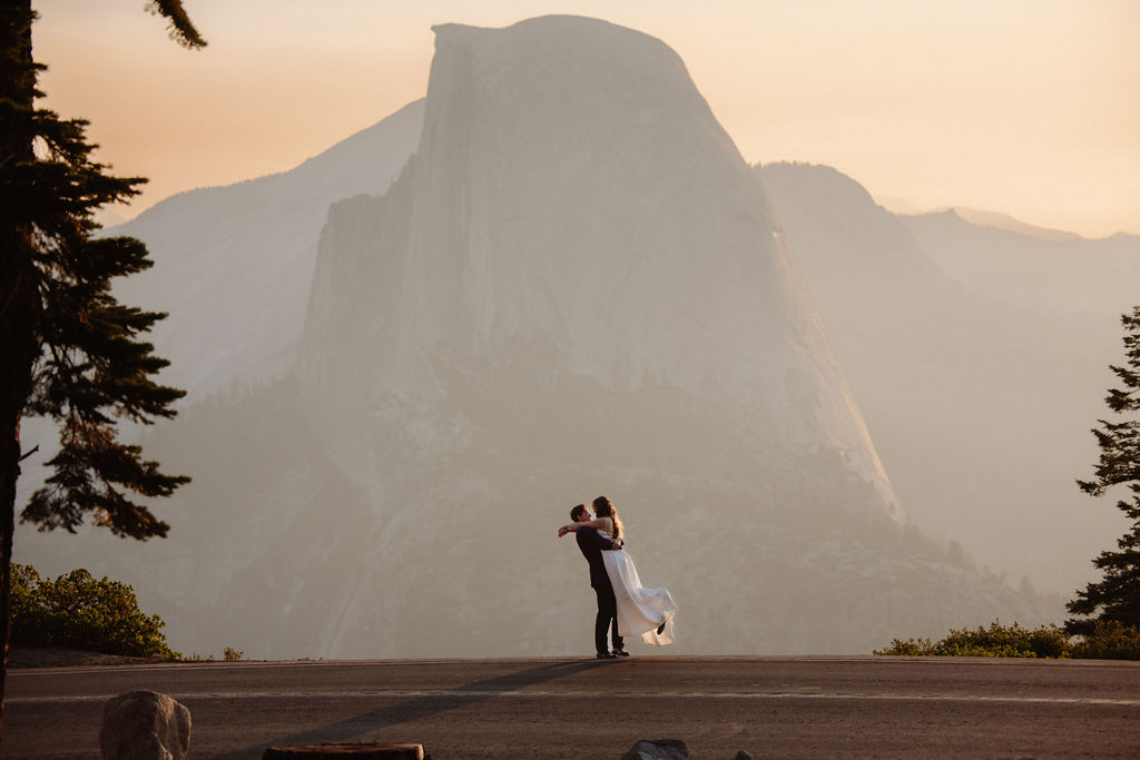 A bride in a white dress stands beside a groom in a suit on a rocky ledge, with mountains and trees in the background for a wedding at glacier point