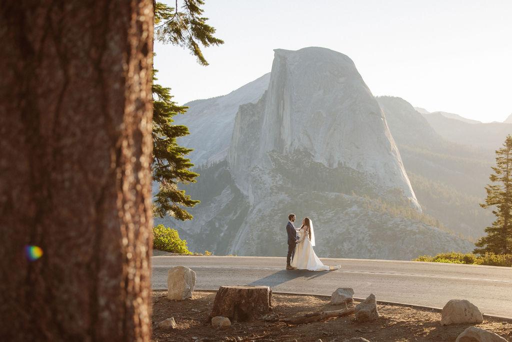 A bride and groom embrace outdoors with mountains and trees in the background, including a prominent granite peak for a wedding at yosemite