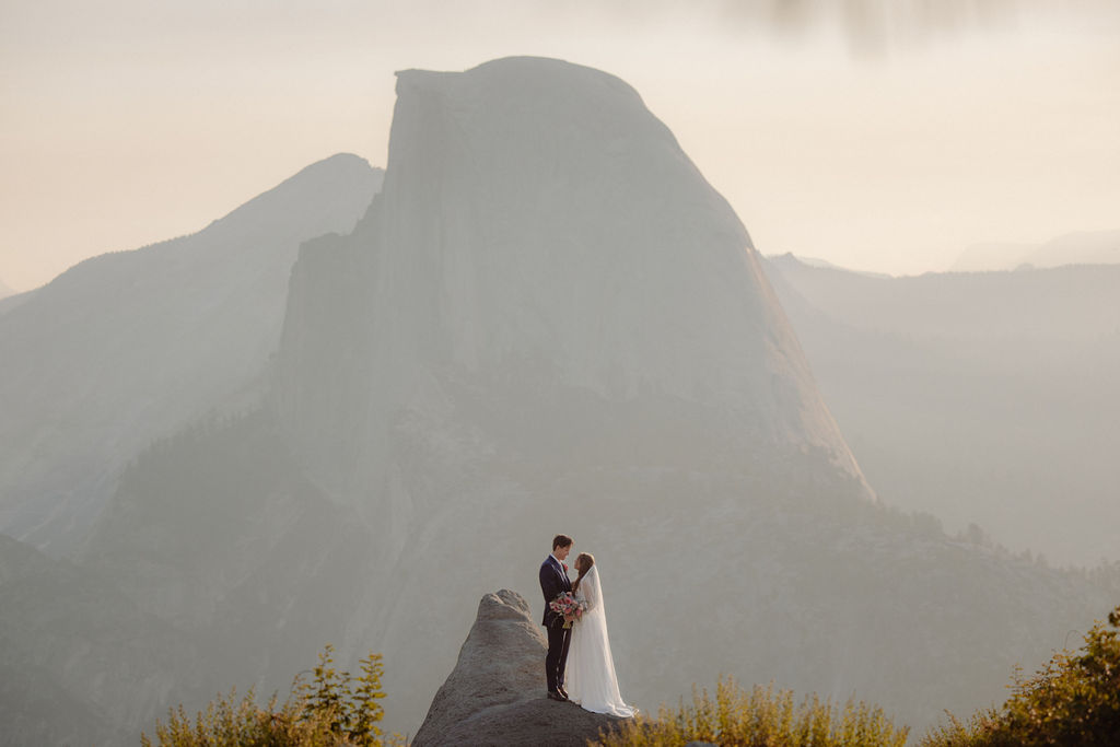 A bride in a white dress stands beside a groom in a suit on a rocky ledge, with mountains and trees in the background for a wedding at glacier point