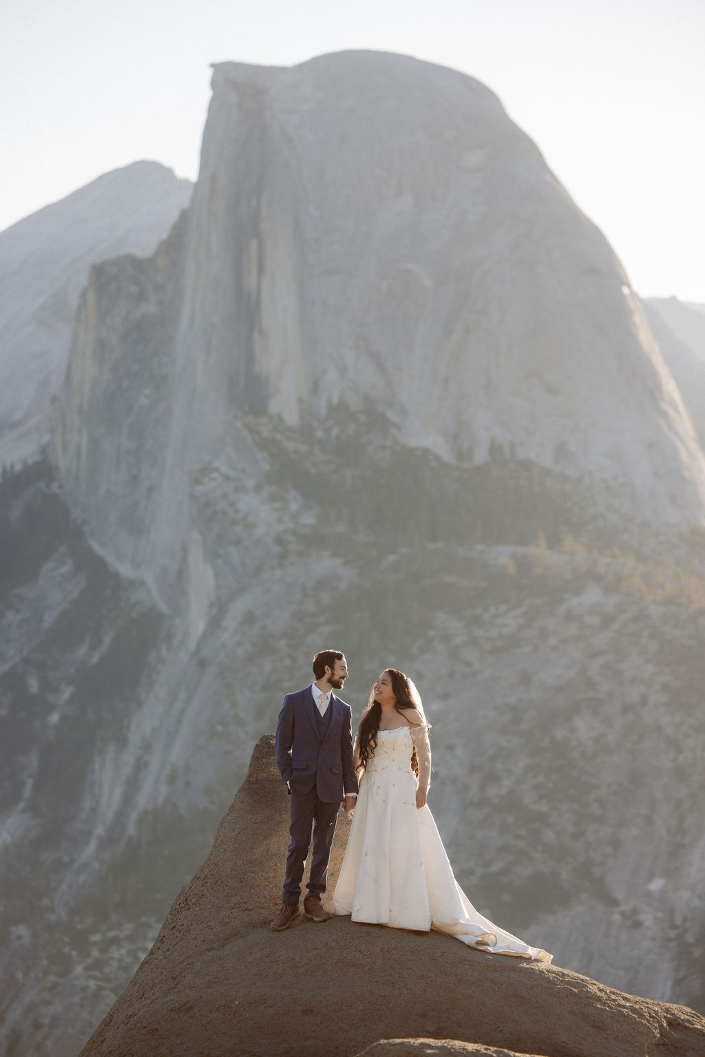 A bride and groom stand together on a rocky ledge with a large mountain peak in the background for a wedding at yosemite