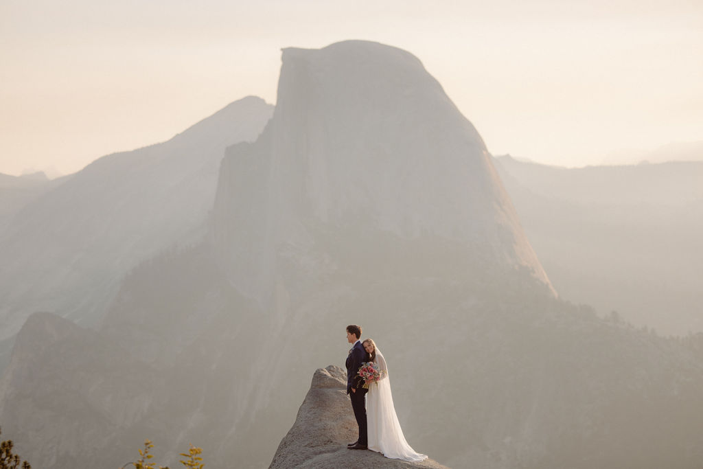 A bride in a white dress stands beside a groom in a suit on a rocky ledge, with mountains and trees in the background for a wedding at glacier point